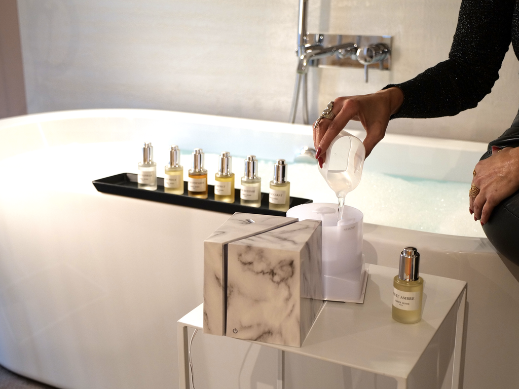 une personne mettant de l'huile de parfum ambre home dans le diffuseur marbre blanc près d'une baignoire 